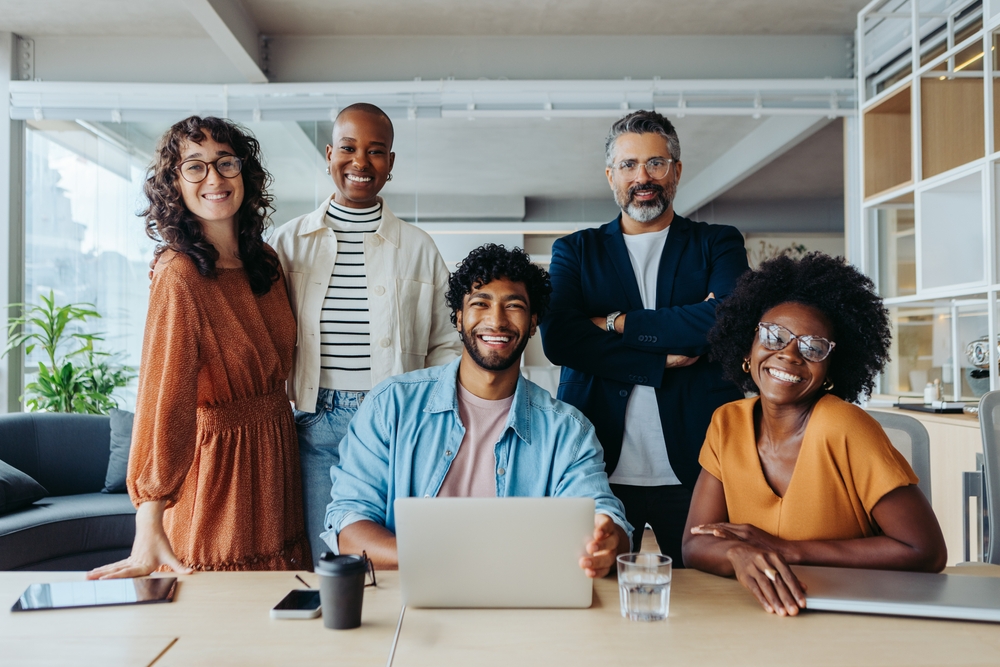 Diverse group of professionals posing together in an office with a laptop on the table.